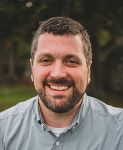 Matt Miller smiling with short graying dark brown hair, mustache and beard and wearing a button up shirt with small blue and white check pattern