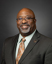 headshot photo of Michael Henry with a black and gray mustache and beard wearing glasses and a dark gray suit with a light blue button down shirt and gold tie