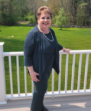 Sharon Mutrie standing on a deck smiling with short brown hair and wearing blue pants and top. Her hand is on the railing and a green lawn surrounded by trees is in background.