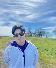 Desmond Ng with short dark hair and sunglasses wearing a light blue zip up sweatshirt and standing on a hill with trees at the top and a blue sky with wispy clouds in the background