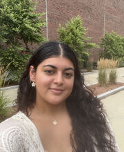 Gabriela Perreira smiling with long dark hair, wearing a white top, standing in front of a brick building with trees beside it