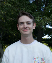 Joey Sebby smiling with short brown hair and wearing a white jersey. There are trees in the background.