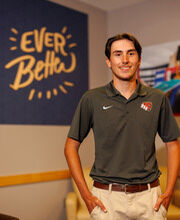 Joshua Nichols standing and smiling with short dark brown hair in front of a BSU Ever Better sign. He is wearing a dark gray BSU polo shirt and khakis.