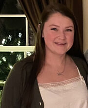 Kara Aries smiling with long dark brown hair, wearing an open brown sweater over a white blouse
