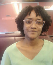 Katia Lopes smiling with medium length black curly hair and wearing clear rim glasses and a light blue green v-neck top