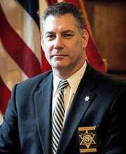 Peter Koutoujian with short styled salt and pepper colored hair wearing a navy blue blazer over a white button down shirt and navy and white striped tie. He is also wearing a badge over his breast pocket.