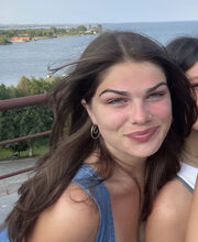 Sam Oldham smiling with long dark brown hair and wearing a denim sleeveless top in front of a railing with the ocean in the background