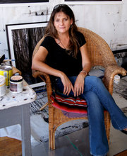 Mary Dondero sitting on a rattan chair in her studio with a framed picture on the floor behind her and a table with paint cans and brushes on it next to her