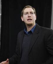 Blake Ruehrwein speaking or teaching wearing a black suit coat over a dark gray v-neck sweater over a blue collared button down shirt