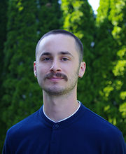 Headshot photo of Professor Eric Aldieri with very short brown hair and mustache wearing navy blue jersey with white trim collar in front of tall green bushes