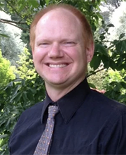 Jon Amon smiling with receding short red hair wearing navy blue button down shirt with gray blue tie and trees in background
