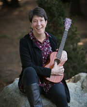 Paula Bishop sitting on a rock smiling with short dark brown hair, holding a ukulele and wearing black leggings and tall fashion boots and a black open sweater over a long maroon button up blouse