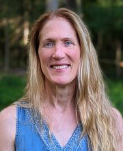 Dr. Darcy Boellstorf smiling with long straight dirty blond hair wearing a v-neck sleeveless blue top with grass and trees in photo background