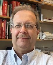 Dr. Ed Brush smiling with short light brown/gray hair and mustache and wearing clear rim glasses and pinstripe white and blue button down shirt with office bookshelves in background