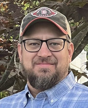 Emmett Buhmann with short gray brown hair, mustache and beard wearing black rim glasses a green and khaki baseball cap and a blue and white plaid button down shirt