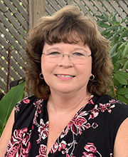 headshot photo of Dr. Andrea Cayson with medium length brown wavy hair wearing clear rimmed glasses and a sleeveless v-neck black blouse with pink floral print