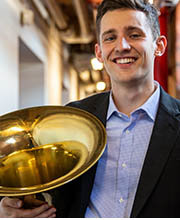 Austin Comerford wearing a black blazer over a blue and white check button down shirt and holding a tuba in his arm