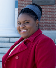 Daniela Belice standing on the steps of Boyden Hall smiling at the camera with her black hair pulled up in a bun and wearing a red coat with lapel and buttons