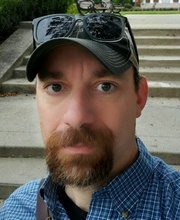 headshot photo of Dr. Giampaolo Digragorio with short dark brown hair and reddish brown mustache beard wearing a black baseball cap with black rim sunglasses on top and a blue checkered button up shirt