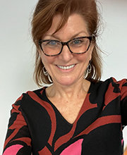 Headshot photo of Dawn DiMartino smiling with shoulder length red-brown hair wearing brown rim glasses and a black, maroon and pink v-neck long sleeve top