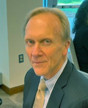 headshot photo of Professor Tom Dunse with short gray hair wearing a brown suit with light blue button down shirt and a tan tie