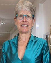 Dr. Susan Eliason smiling with short gray hair and wearing teal blue rim glasses and a teal blue wrap dress with collar