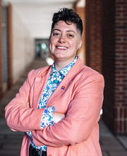 Dr. Reihonna Frost-Calhoun smiling with short dark brown curly hair wearing a salmon colored blazer with rainbow flag pin on it and blue and white print button down shirt under it