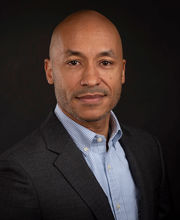 headshot of Dr. Jakari Griffith smiling with no hair and black and gray stubble mustache and beard wearing a blue and white pinstripe button down shirt under a dark gray suit jacket