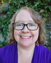 Dr. Ashey Hansen-Brown smiling with medium length wavy light brown hair and wearing brown rim glasses and a royal blue v-neck top
