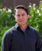 Dr. Asa Iacobucci standing outside in front of greenery with short brown hair wearing a black and purple check button up shirt