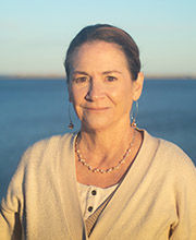 headshot photo of Dr. Margaret Kjelgard with her brown hair pulled back, smiling and wearing a beige v-neck sweater with ocean in the background
