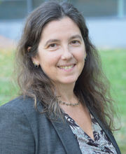 Brenda Kozuch smiling with dark brown wavy long hair wearing a charcoal gray blazer over a v-neck gray and black top