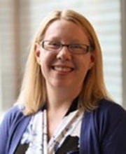 Dr. Heather Marella smiling with medium length blonde hair wearing brown rim glasses and a vneck black white and purple print dress with a purple jacket