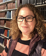headshot photo of Linda Meiberg in front of library bookshelves with light brown medium length curly hair wearing black rim glasses and a black and salmon top under a black vest