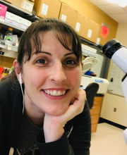 Dr. Joslyn Mills next to microscope in lab smiling with long dark brown hair pulled back in a pony tail with bangs and wearing earphones and a black long sleeve top