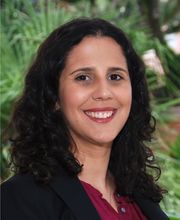 headshot photo of Dr. Rachel Navarre smiling with long dark brown curly hair, wearing a black blazer over a maroon button down top