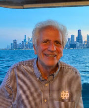 Leonard Paolillo on a boat smiling with short gray hair wearing a black and white checked button down shirt with water and a city skyline in the background