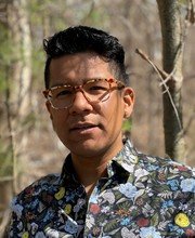 headshot photo of Dr. Luis Paredes with short wavy dark hair, wearing light brown glasses and a floral button down shirt with a dark background. Woods are 