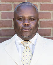 headshot photo of Pierre-Louis Richardson with graying short black hair and goatee wearing a cream colored blazer over a white button down shirt with thin blue stripes and a pale yellow tie with small blue squares on it