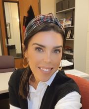 Dr. Ashley Rodrigues smiling with straight brown hair pulled back with a headband wearing a black knit vest over a white button-down blouse