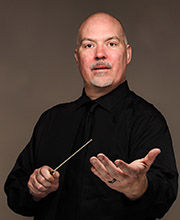 Dr. Donald Running wearing a black button down shirt with black tie, holding a conductor's wand in one hand while his other hand is held out towards the camera