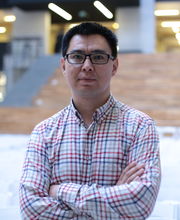 Dr. Rahat Sabrybekov with short dark hair wearing black rim glasses and a red, blue, green and white checkered button down shirt and standing with his arms folded in front of him
