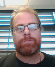 Dr. John F. Santore with reddish blonde receding hair, mustache and beard wearing brown rim glasses and a dark green shirt