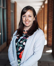 Courtney Sutcliffe smiling with long brown hair wearing a long light blue sweater over a black, blue and coral print top