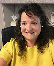 headshot photo of Therese Canuel sitting at her desk smiling with medium length curly dark brown hair wearing a yellow short sleeve top