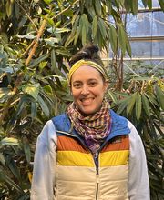 Emma Welch standing in a greenhouse smiling with brown hair in a messy bun on top of her head wearing a gold headband, gray long sleeve sweatshirt over an LLBean puffy vest with blue collar and orange, gold and beige stripes. She also wears 2 fashion scarves in colors of purple, gold and orange