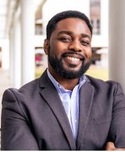 Dr. Maurice Williams Jr. smiling with short black hair, mustache and beard and wearing a light blue button down shirt under a gray suit jacket