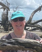 Dr. Catherine Womack on a beach smiling with gray hair pulled back and wearing a teal blue cap with a fish on it, sunglasses, a blue beaded necklace and a black, brown and white patterned short sleeve top. She is holding a large piece of driftwood and there are fallen trees of driftwood behind her and the ocean beyond