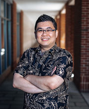 Dr. Cory Yeo smiling with short spiked black and gray hair wearing black rim glasses and a black short sleeve button down shirt with tropical leaves outlined in beige