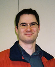 Dr. Michael Zimmerman smiling with short dark brown hair and glasses wearing an orange parka with black collar over a navy blue button down shirt